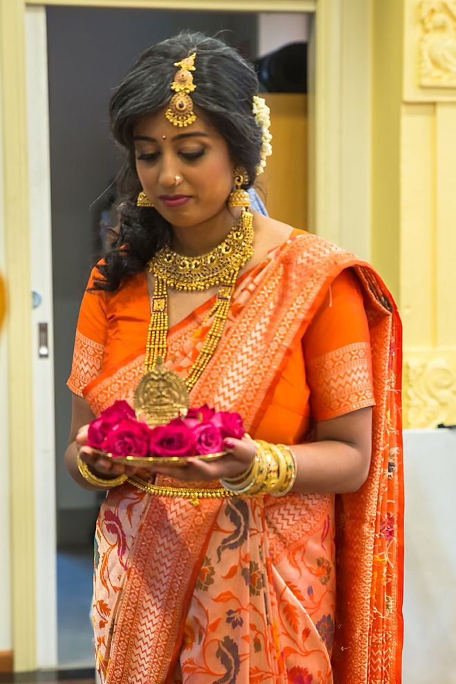 A bride in a vibrant orange and gold traditional Indian wedding sari holds pink roses while getting ready for her ceremony.