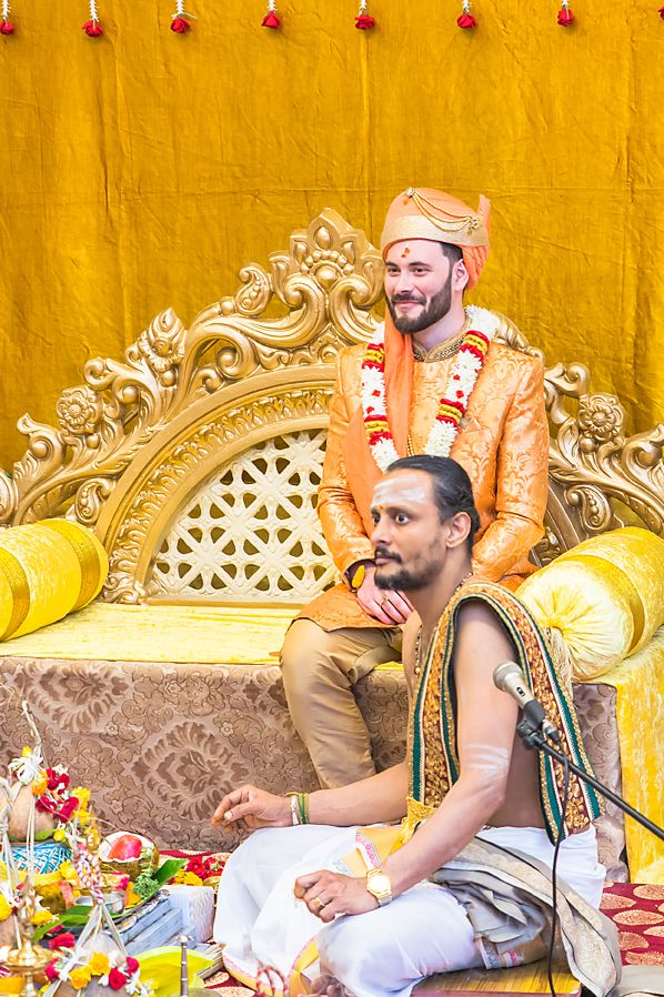 Two men in traditional Indian attire sit on an ornate golden throne against a bright yellow backdrop during a ceremony.