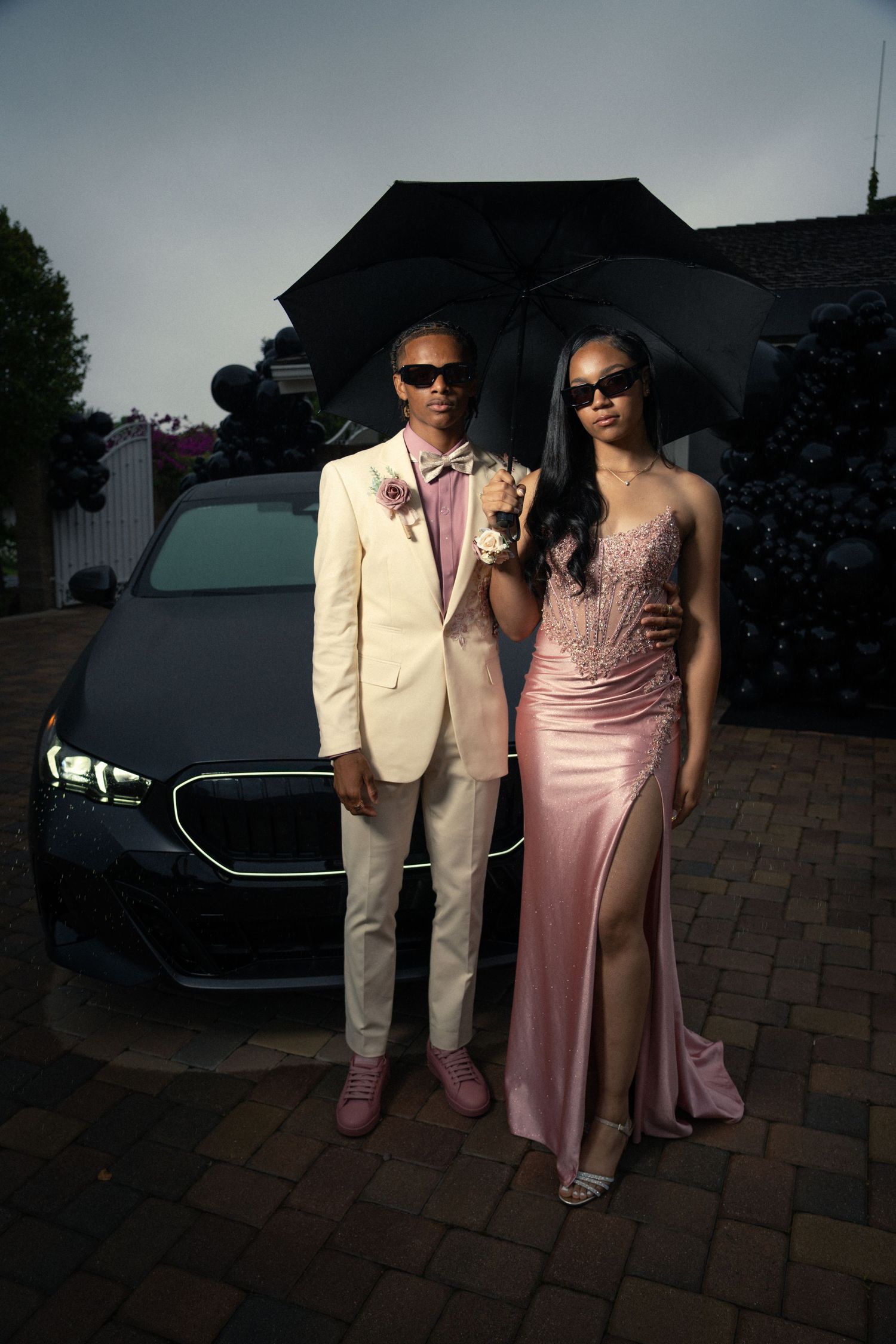 Elegant couple in formal attire poses with umbrellas next to luxury car at dusk.