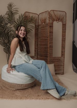 Casual lifestyle photos with white top and blue jeans on round woven ottoman with tropical decor.
