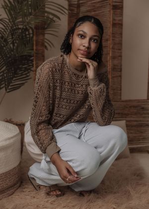 Casual indoor photoshoot with brown knit sweater and white pants against brick wall and palm decor.