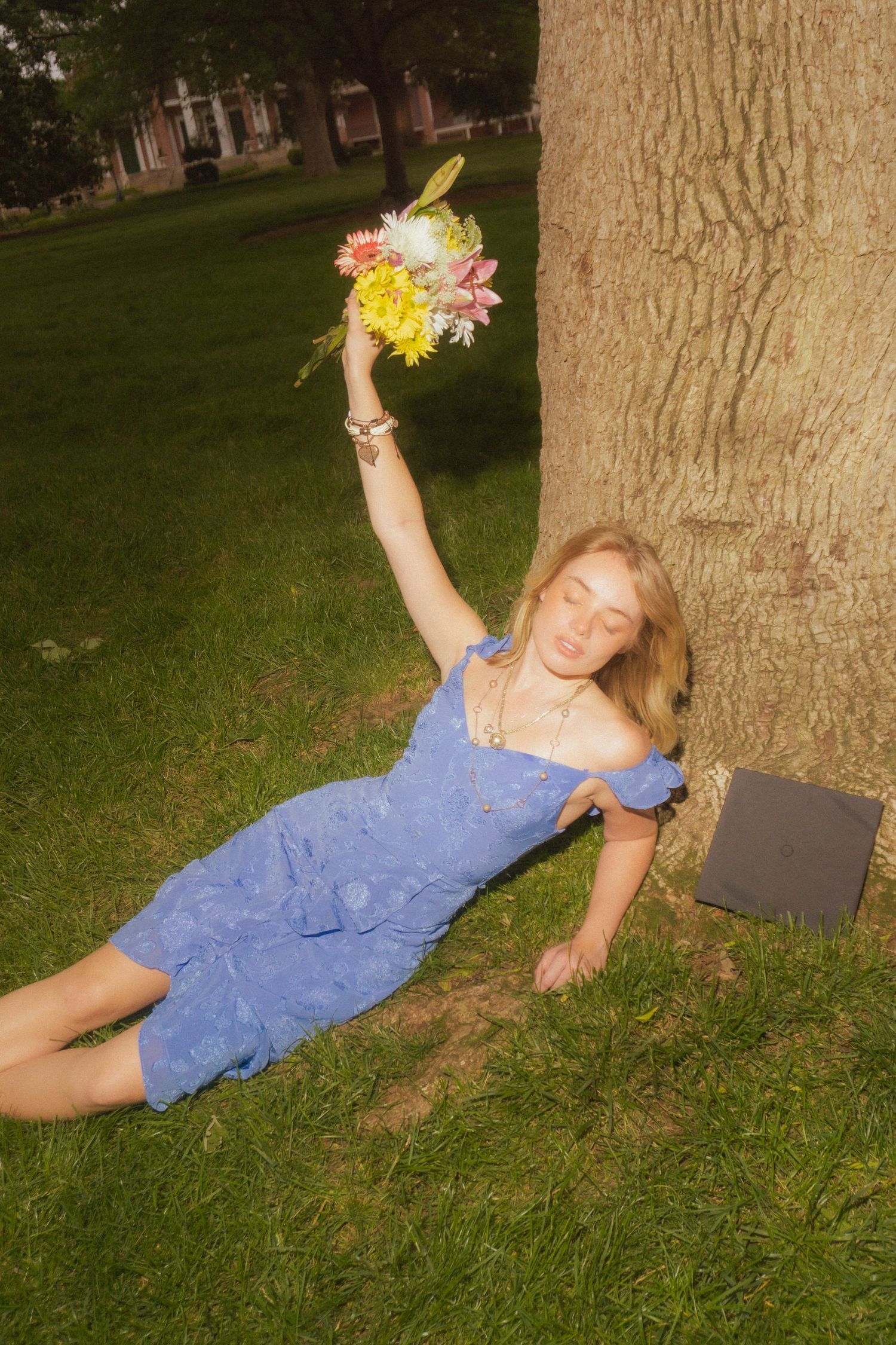 Person in blue dress creates artistic poses against tree trunk while holding flowers.