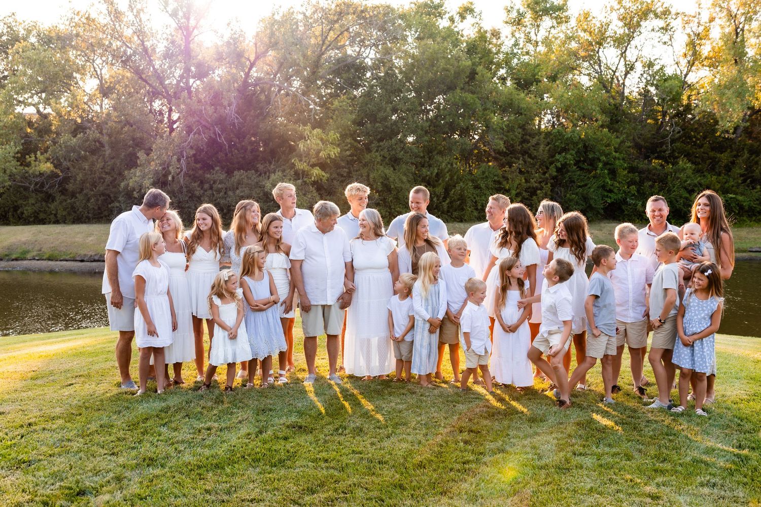 Large group photo of people wearing matching white outfits sitting together in a grassy field at sunset.