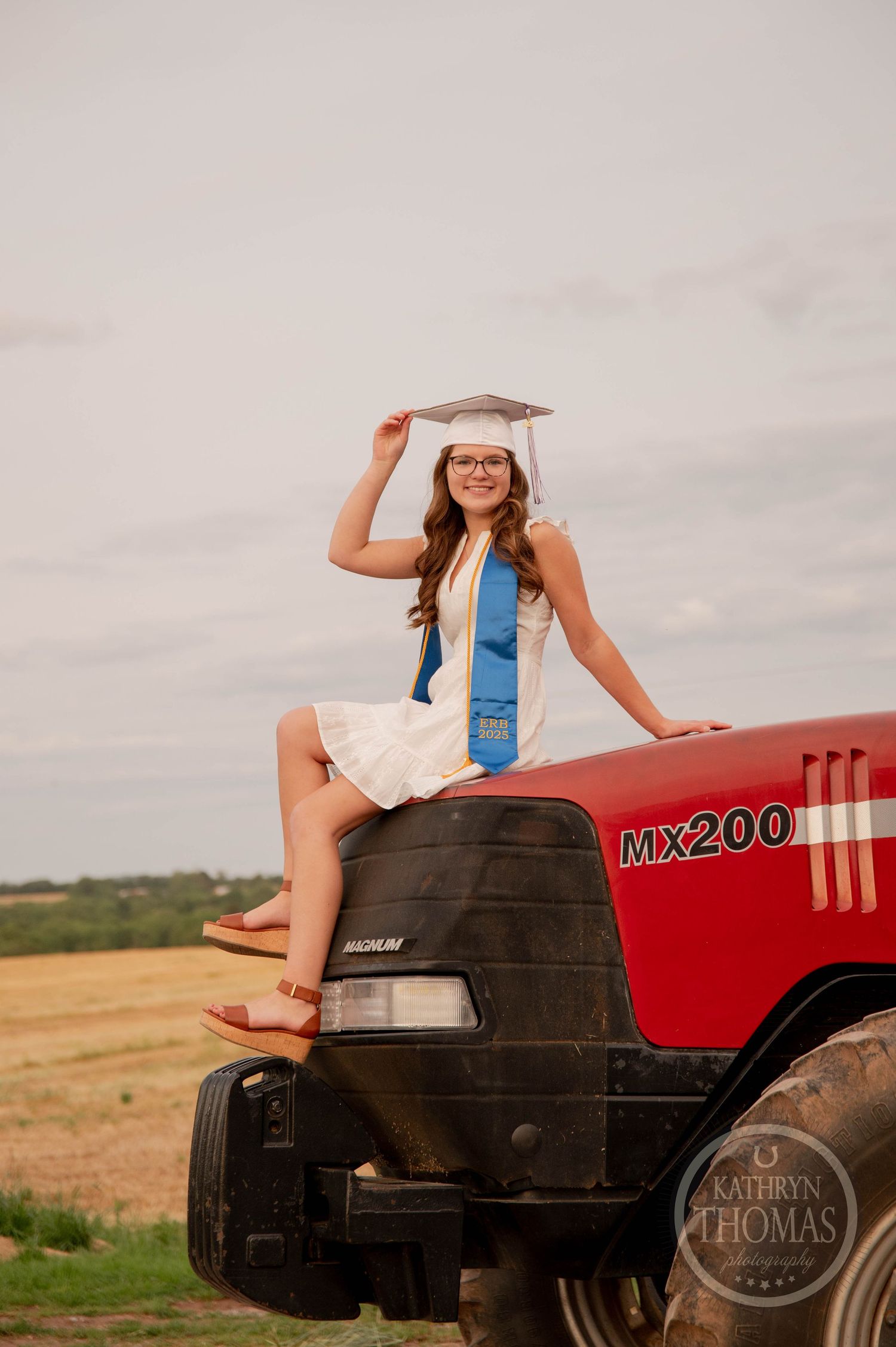 Woman in white dress poses playfully on a red Massey Ferguson tractor in a rural farm field setting.