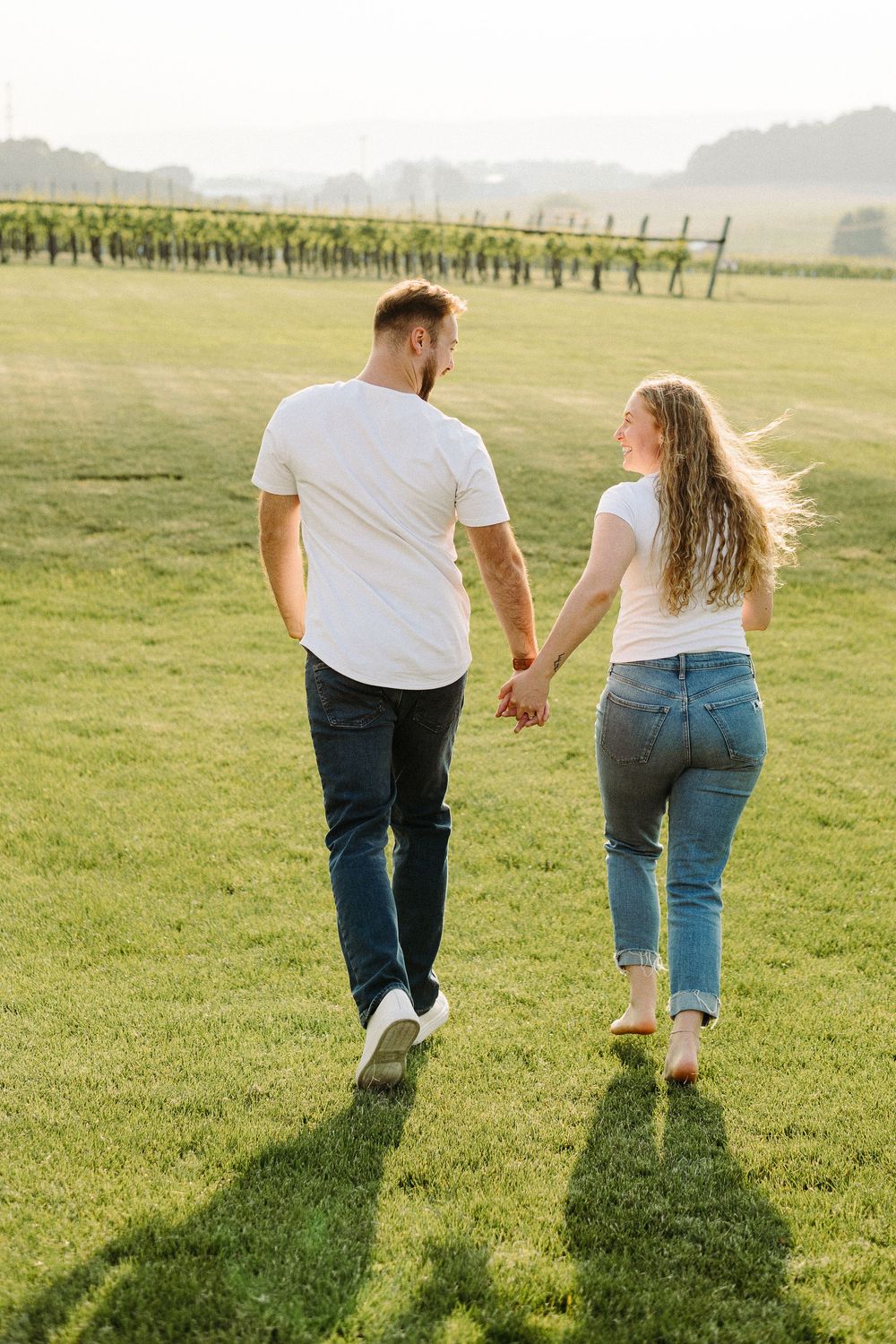 Two people walking hand in hand across a sunlit grassy meadow with distant trees on the horizon.