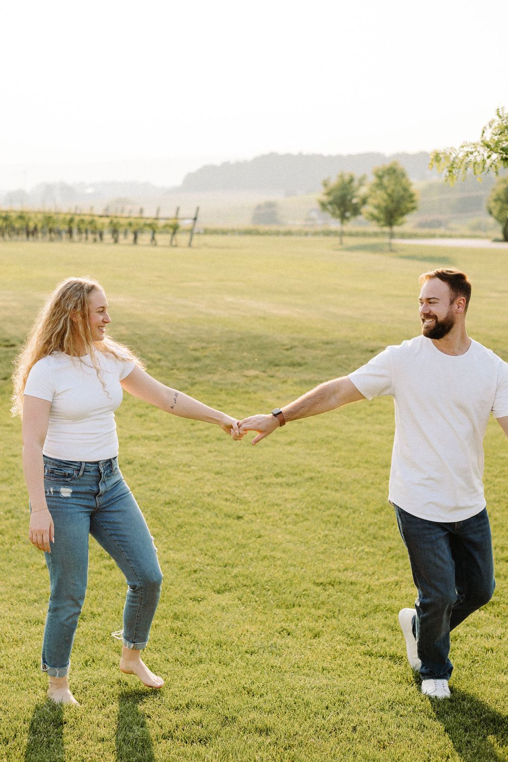 Playful couple in white shirts and jeans dancing together on a grassy meadow during golden hour.