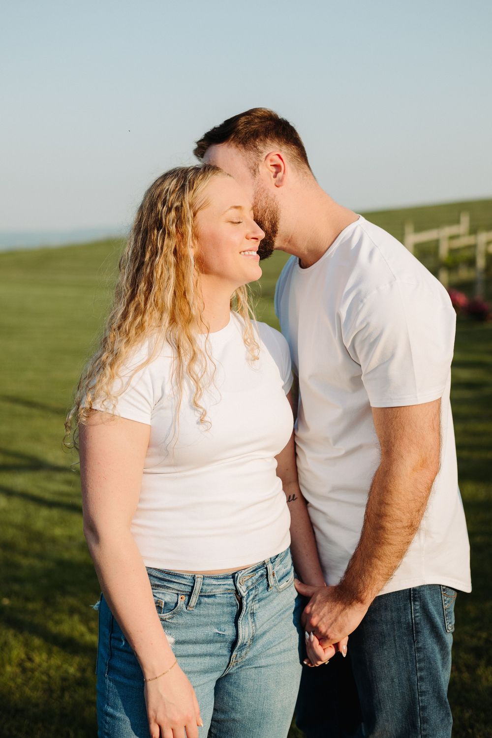 A couple in matching white t-shirts shares a tender moment on rolling green hills at sunset.