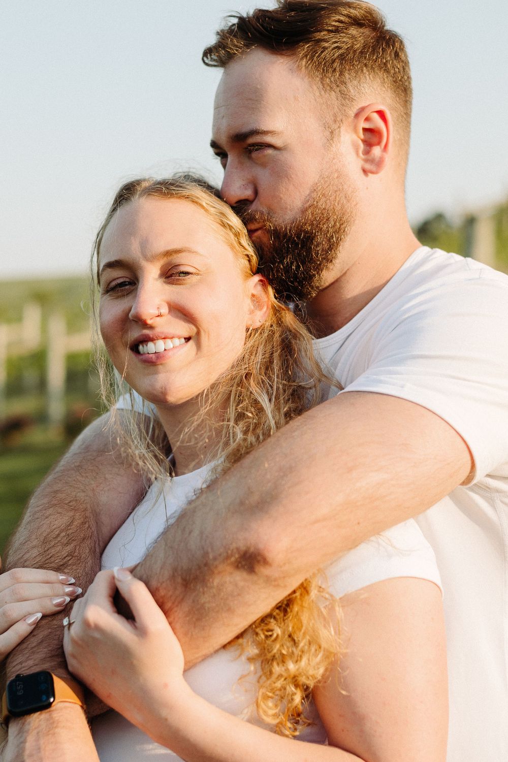 A couple embraces outdoors with warm sunlight, the woman smiling while wearing a white top and blonde curly hair.