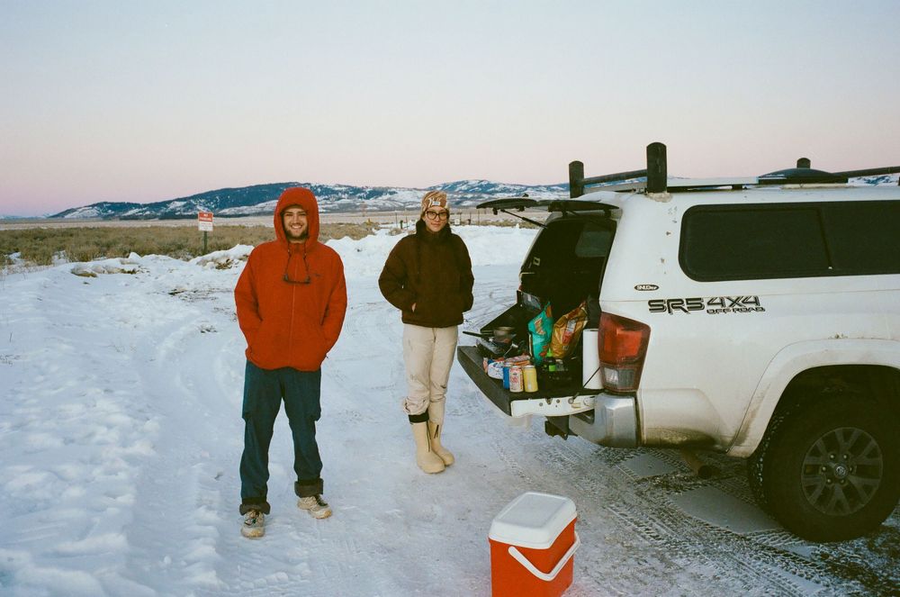 Two people standing in snow beside a white Toyota Tacoma truck at dusk in a winter landscape.