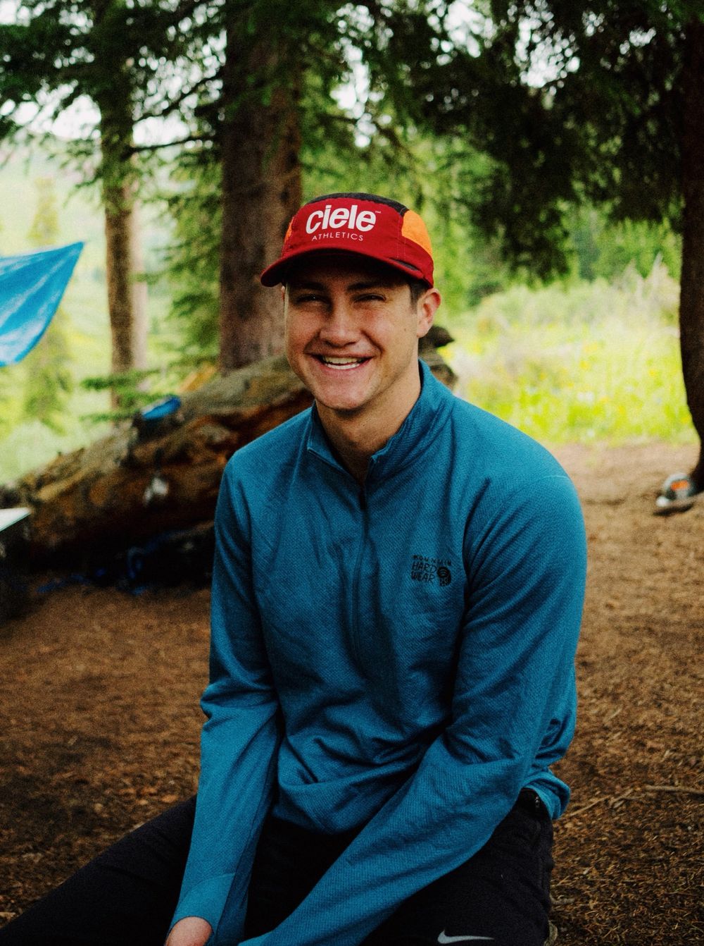 Person wearing blue fleece and red cap sitting outdoors in a wooded setting.
