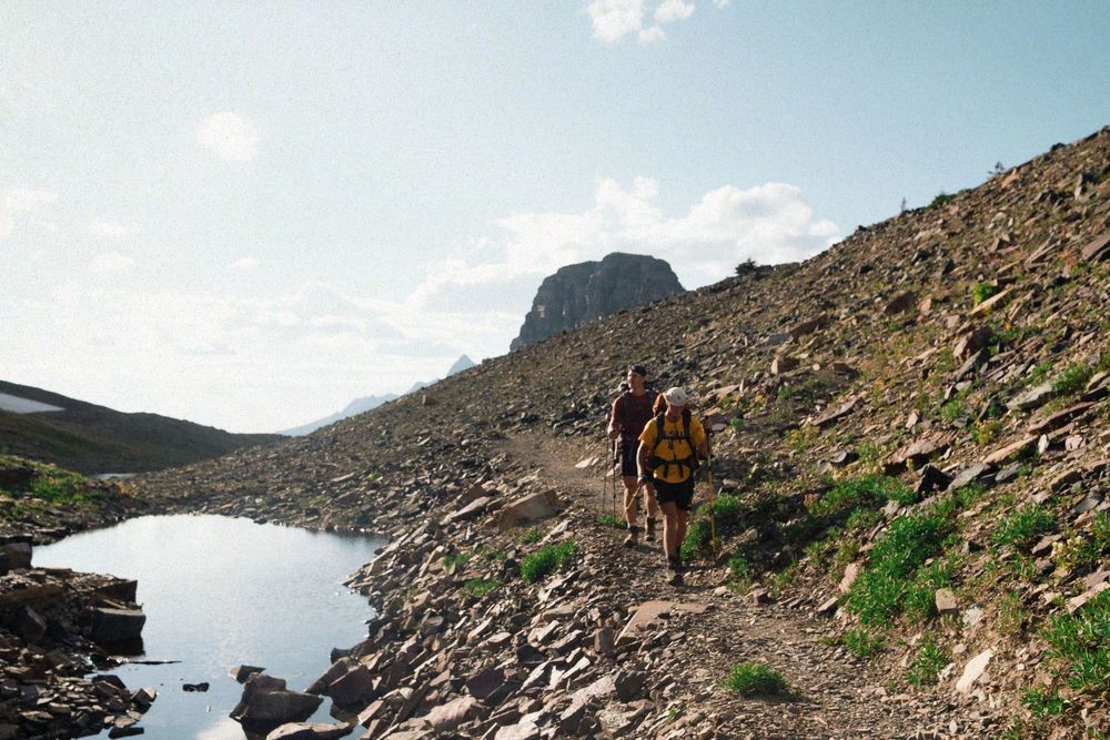 Hiker walking on rocky mountain trail beside alpine lake under blue sky.