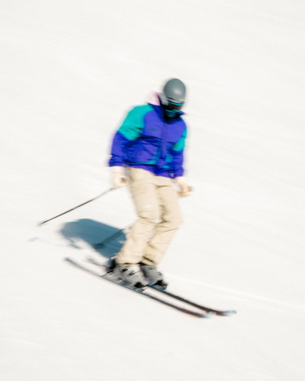 A skier in a purple and teal jacket with beige pants descends a snowy slope, captured with motion blur.