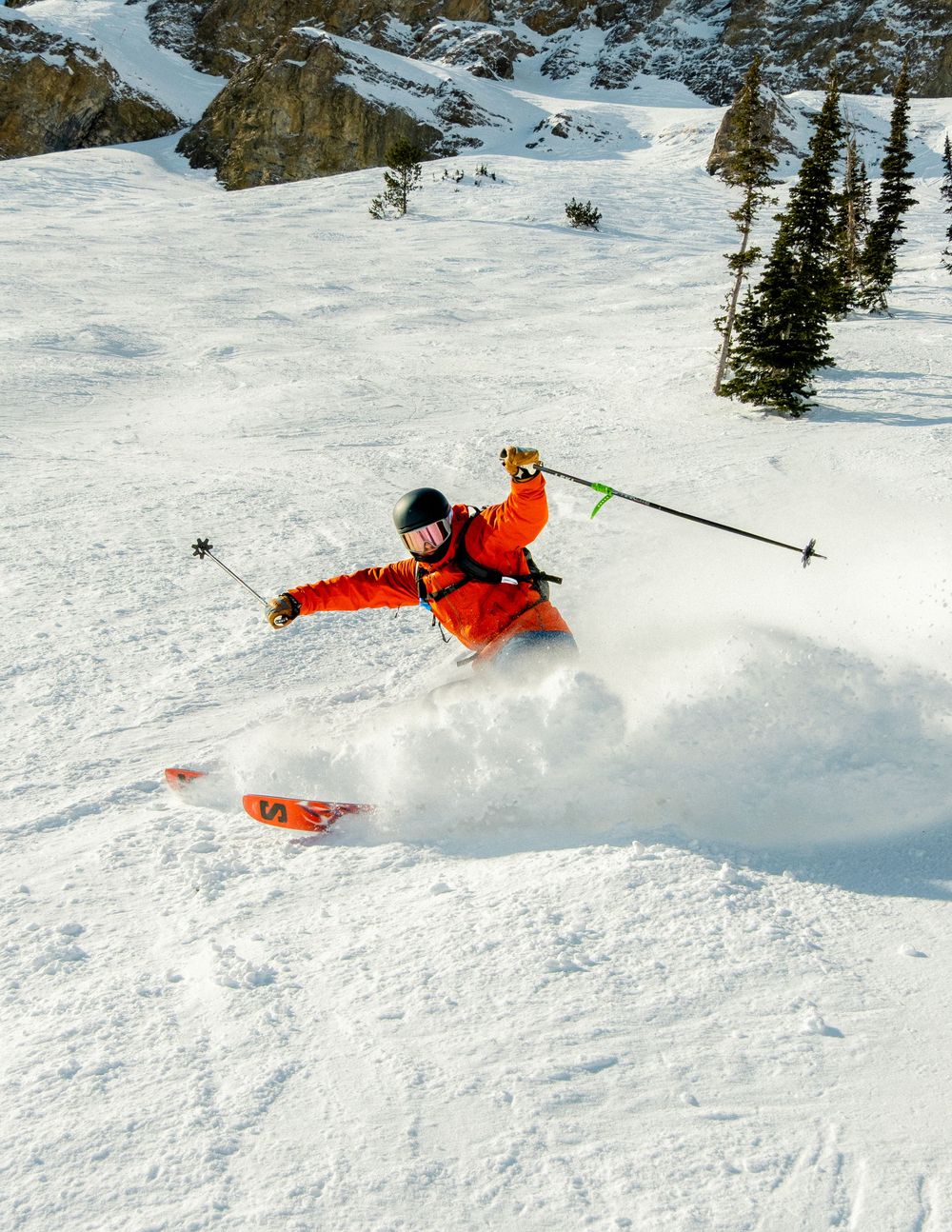 Skier in orange jacket carving through deep powder snow on a steep mountain slope with evergreen trees in background.
