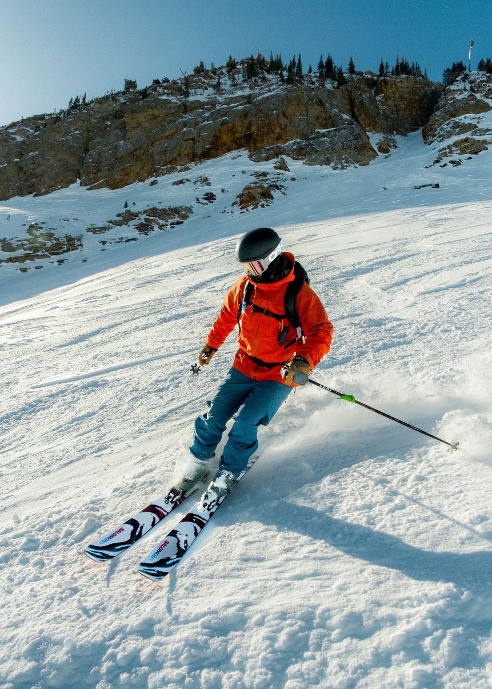 Skier in orange jacket descending snowy mountain slope with rocky cliffs in background on a sunny day.