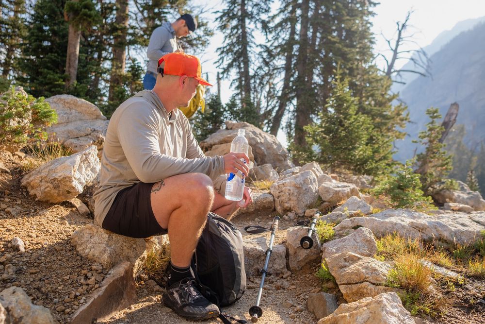 Hiker resting on rocks filtering water with mountains and pine trees in the background on a sunny day.