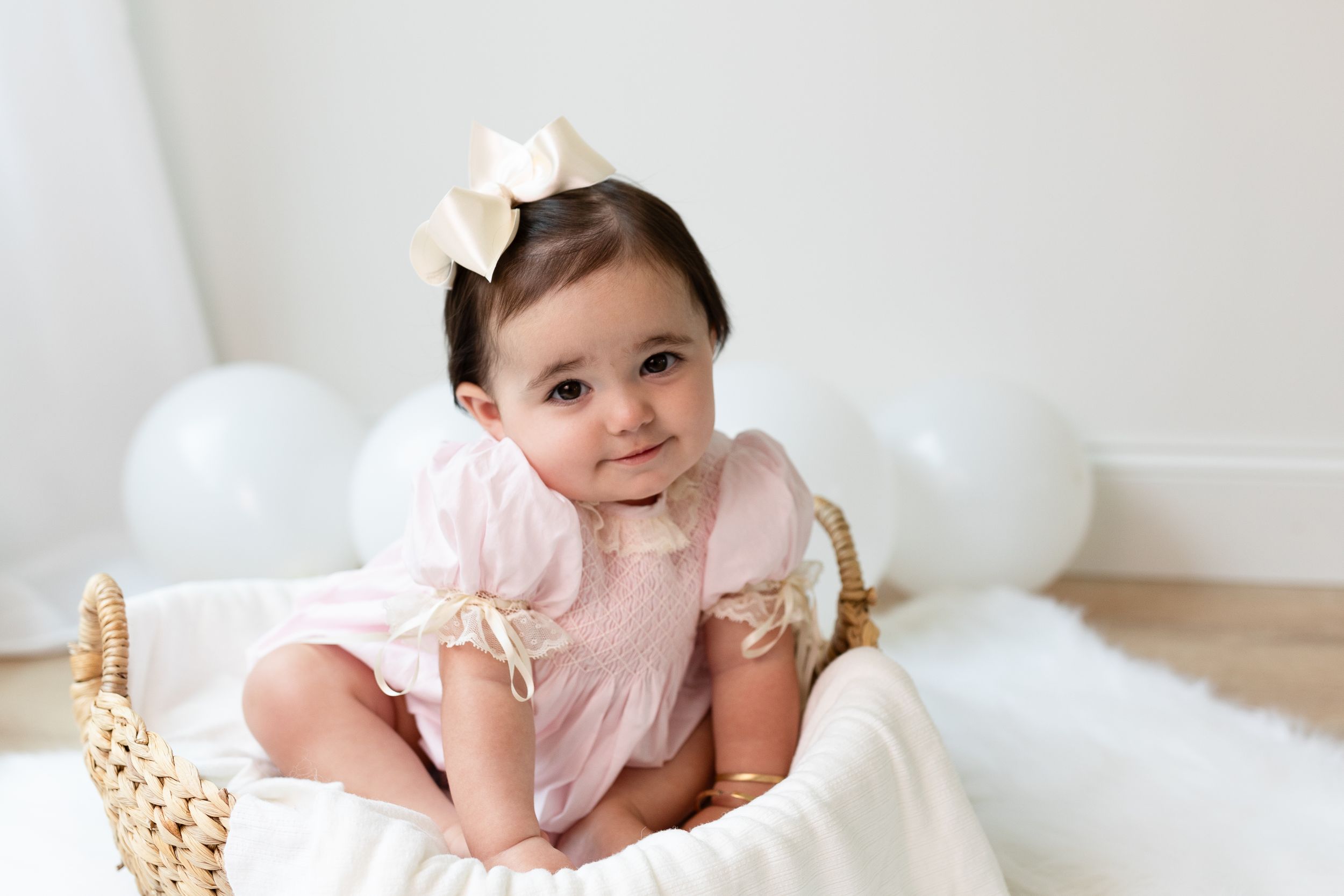 Baby in pink dress with bow sitting on white blanket.