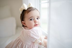 Baby in delicate pink dress near bright window.