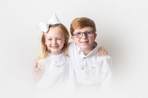 Two children wearing white outfits pose together against white background.