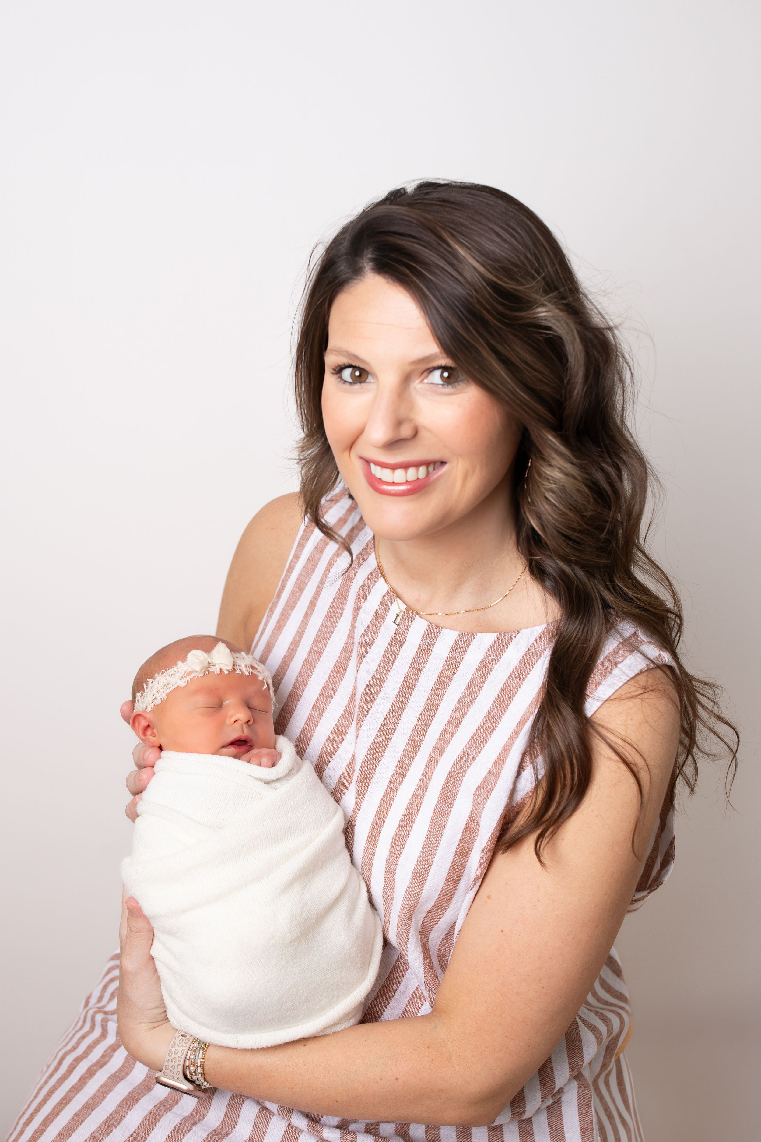 Woman in striped dress holding newborn baby against white studio backdrop.