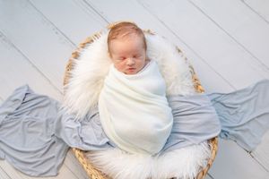 Newborn baby swaddled in white blanket resting on blue fabric with woven basket.