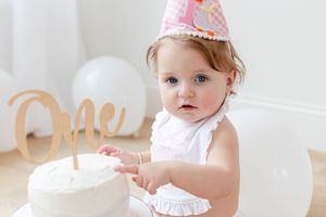 Smiling baby in pink party hat sitting next to white birthday cake on white background.
