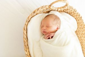 Peaceful sleeping newborn baby wrapped in white swaddle lying in woven basket with natural lighting.
