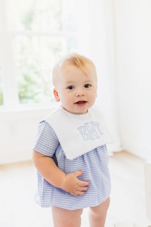Baby wearing blue gingham outfit and white bib smiling at camera.