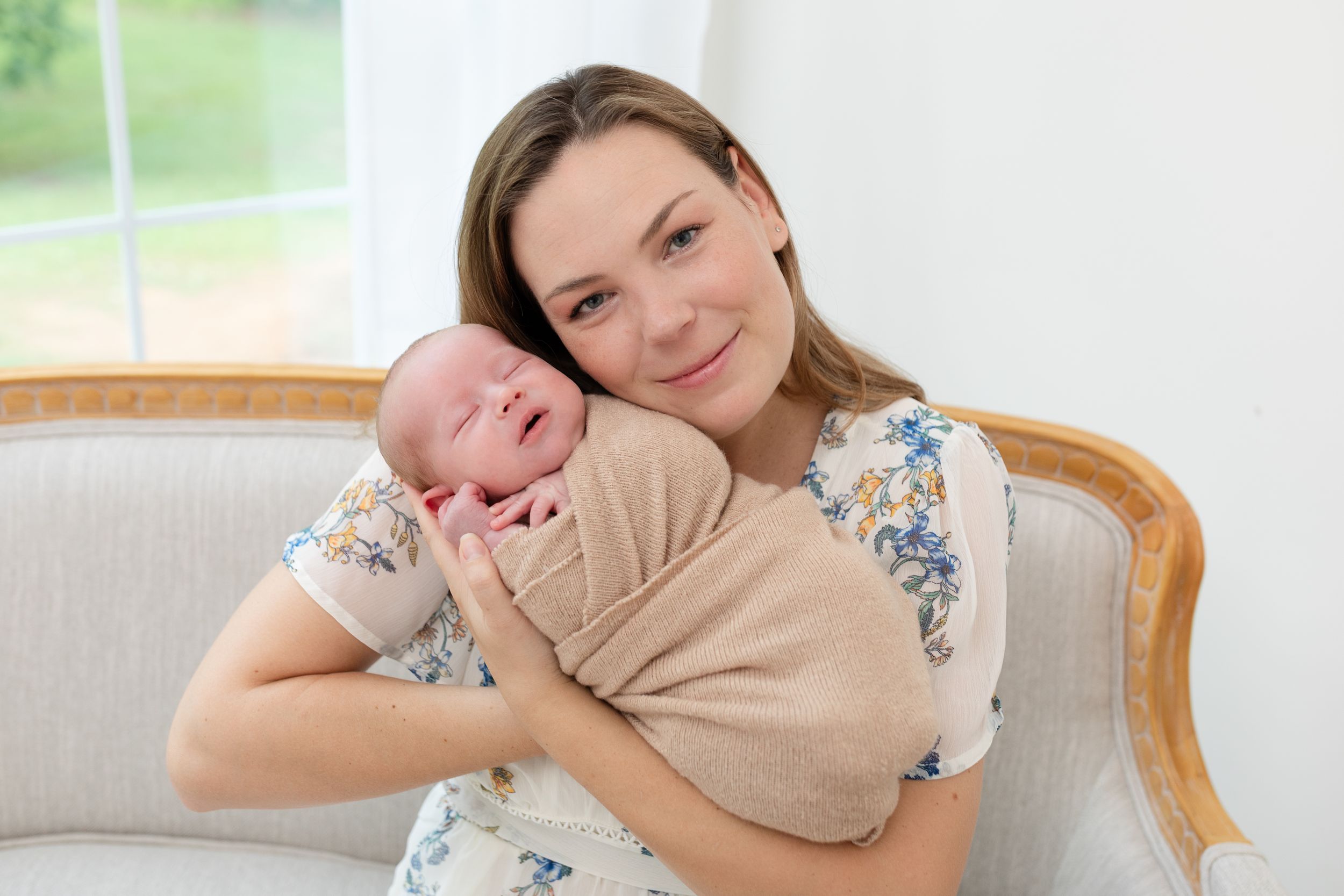 Smiling mother in floral dress holding swaddled newborn baby on a white sofa indoors.