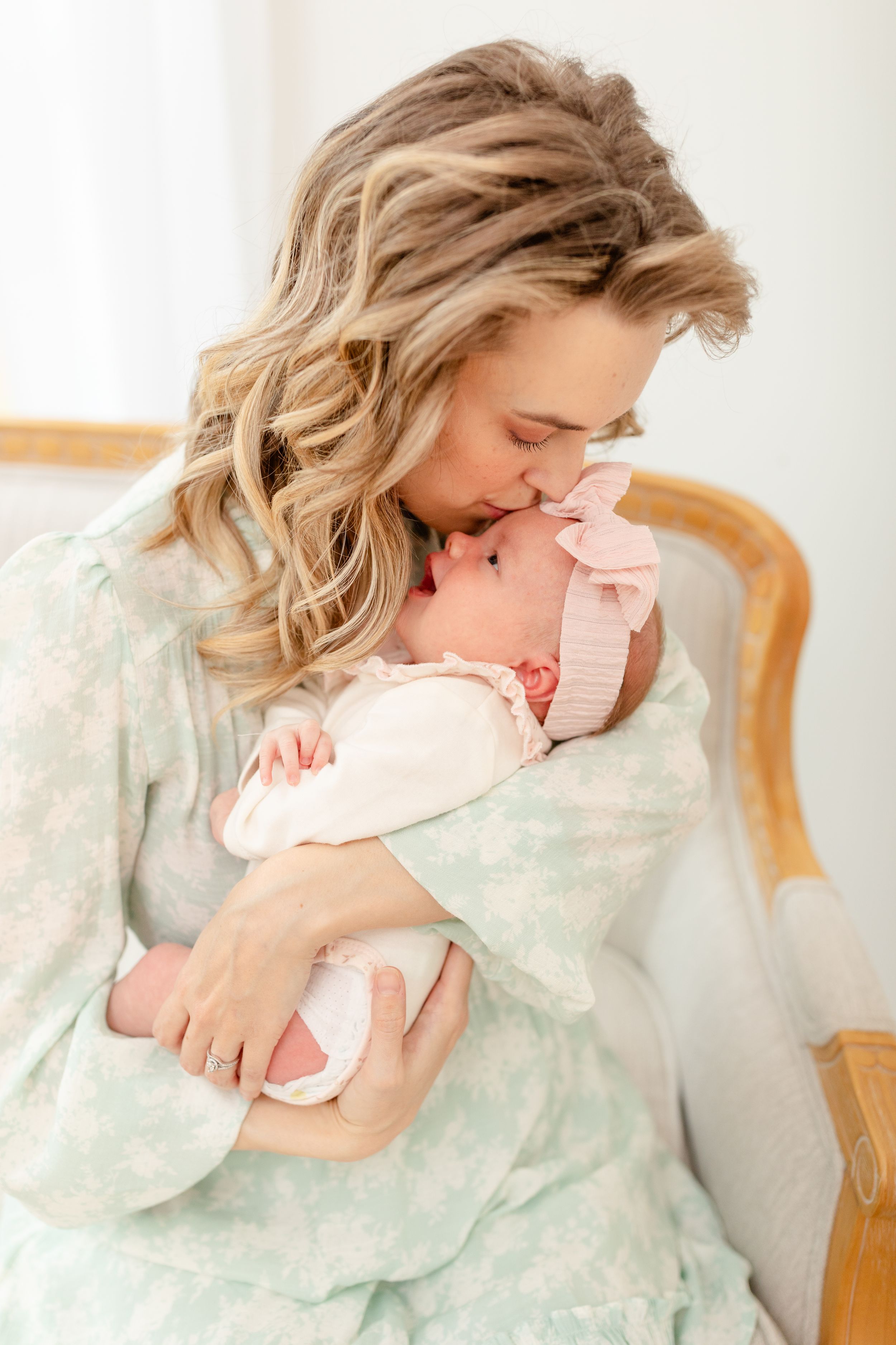 A mother in a gray sweater tenderly embraces and kisses her newborn baby wrapped in a pink blanket.