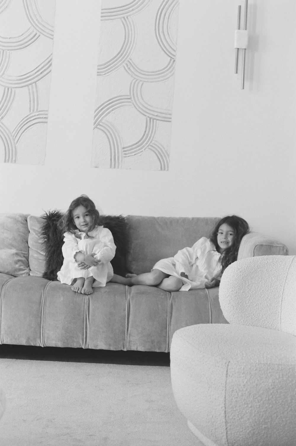 Two children relax on a gray couch in a minimalist room with curved wall patterns and a white accent chair.