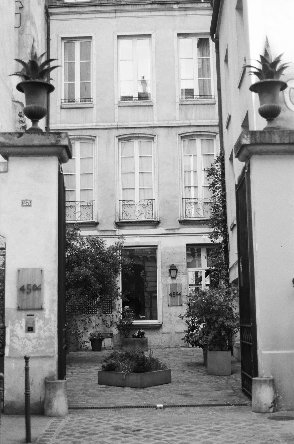 Black and white photo of a quaint Parisian courtyard entrance with potted plants and traditional architecture.