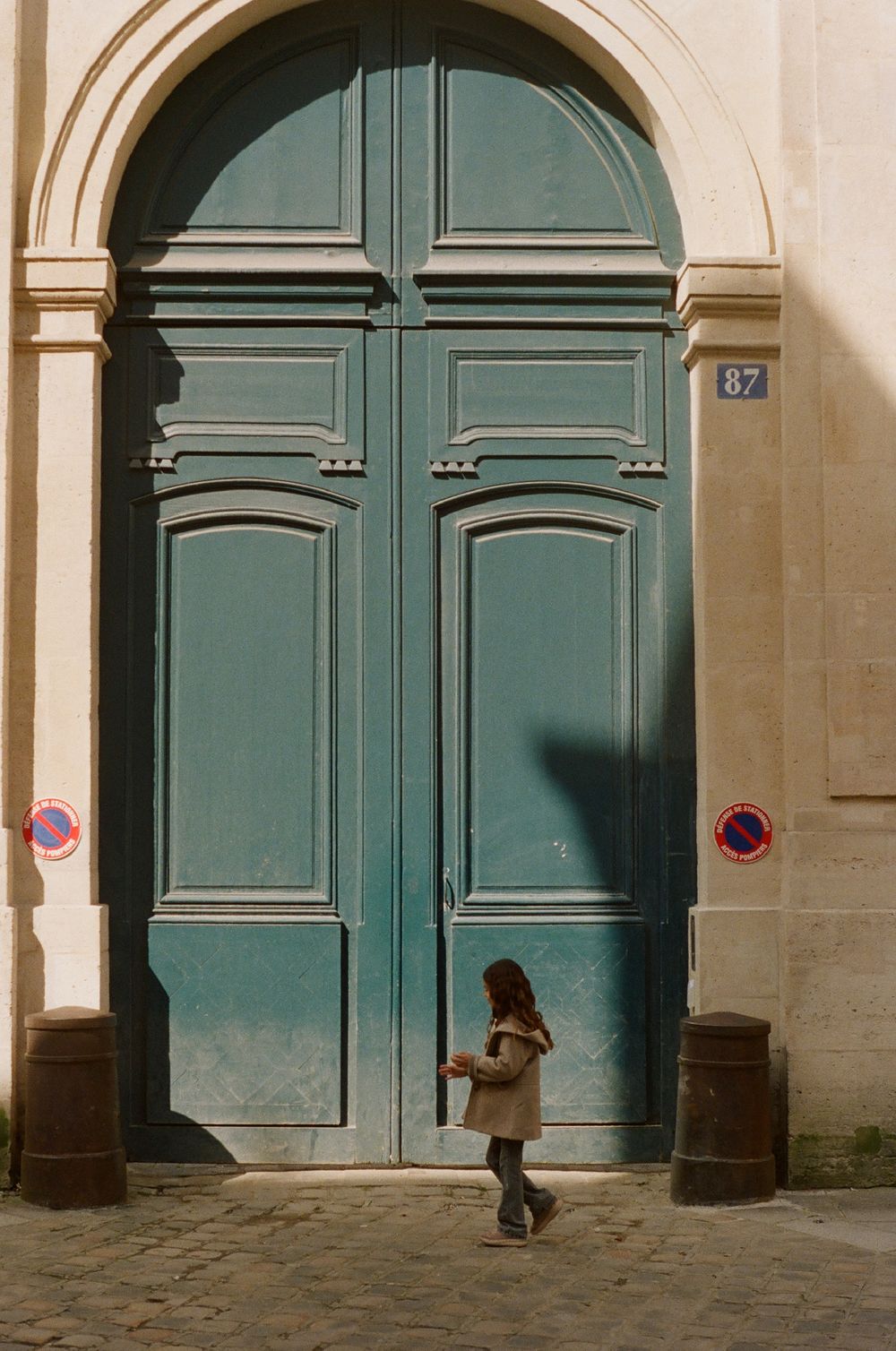 A small figure in a beige coat stands before a large turquoise double door with an arched frame and number 87.
