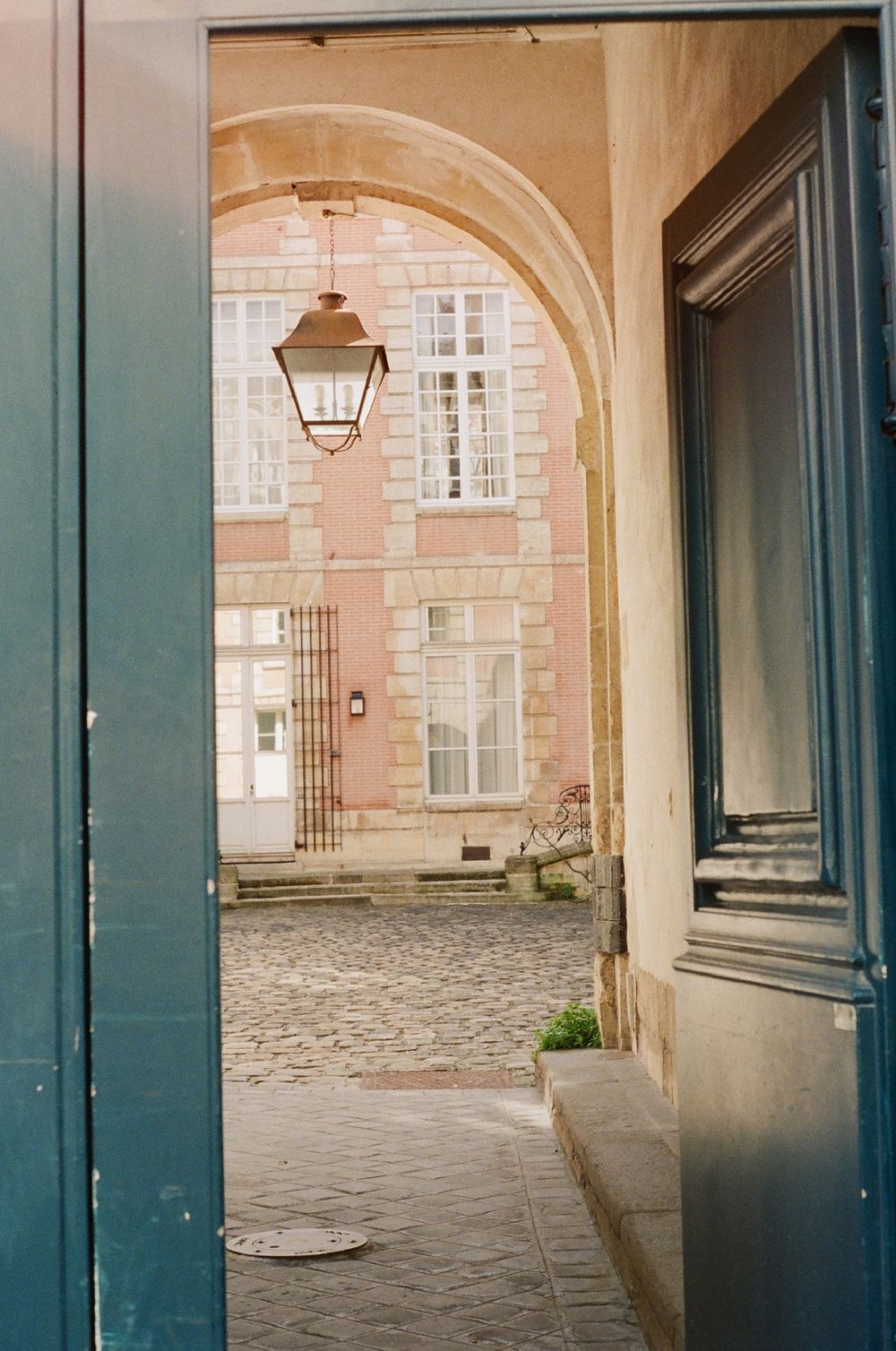 Stone archway entrance with cobblestone courtyard and teal-painted wooden doors in a historic building complex.