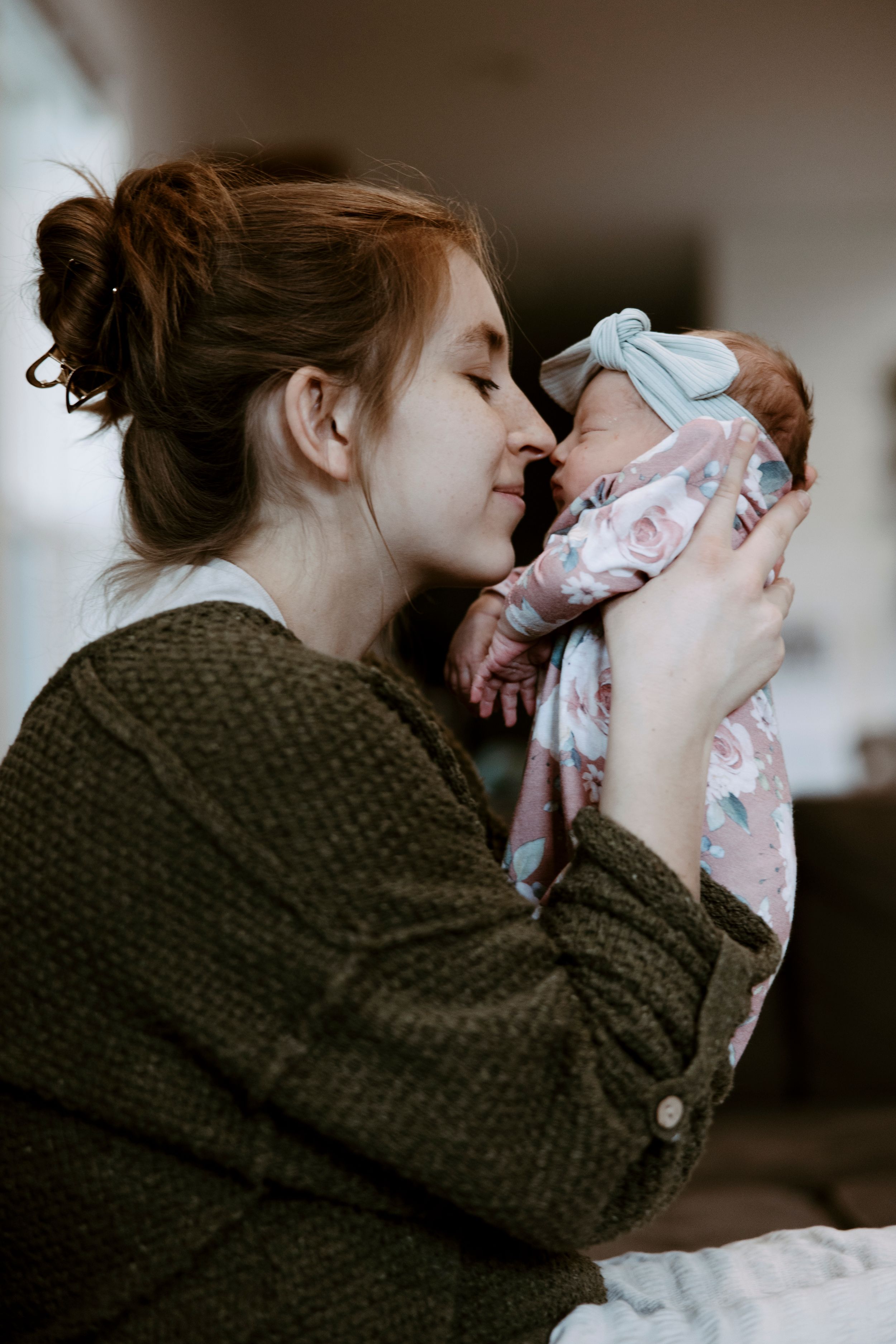 Red-haired mother tenderly nuzzling her newborn baby dressed in floral outfit with blue bow.