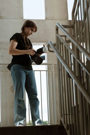 Young woman in black shirt and jeans adjusts camera lens while standing on outdoor concrete staircase.