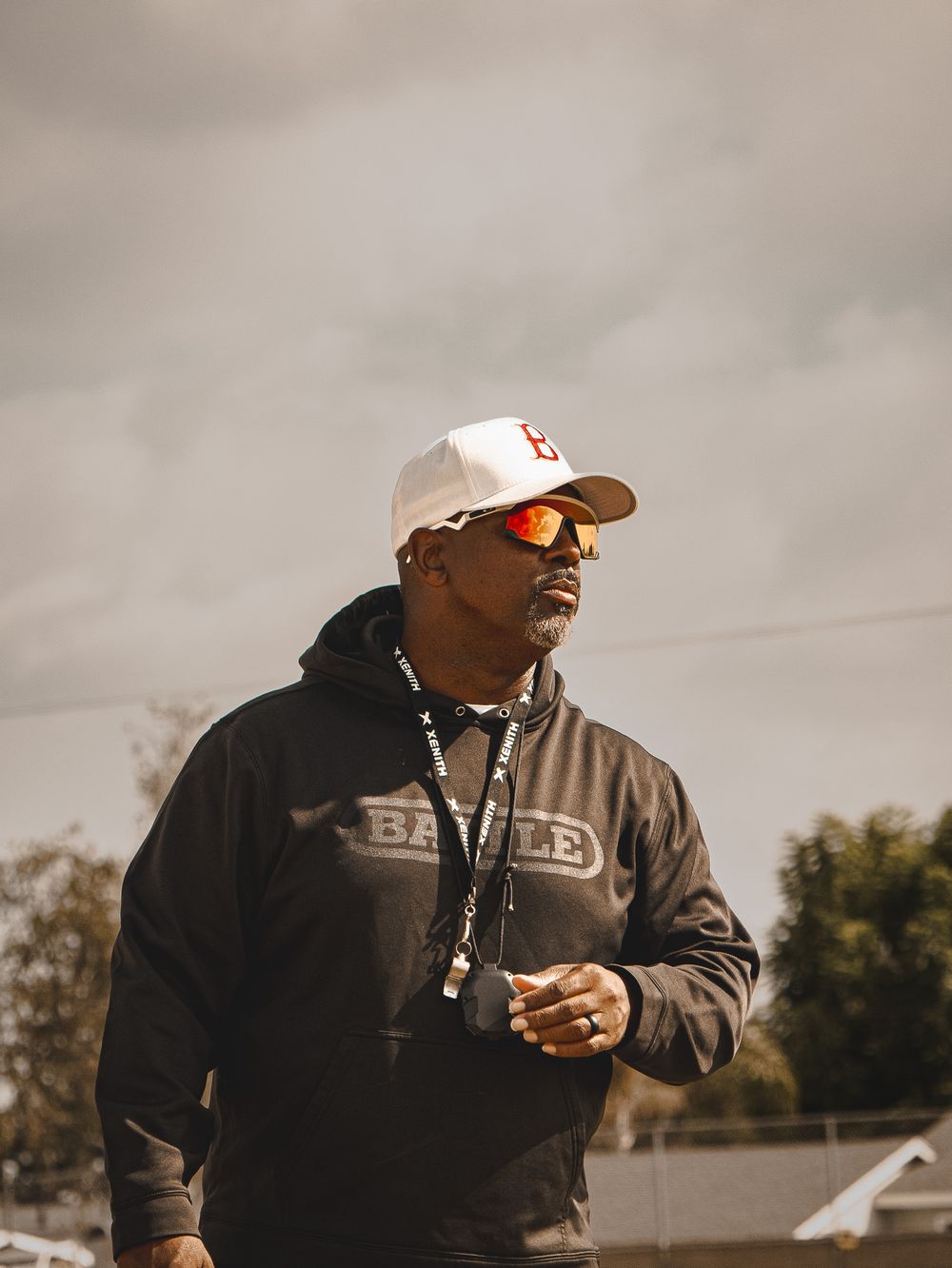 A coach wearing a black hoodie, white cap and sunglasses stands on a sports field.