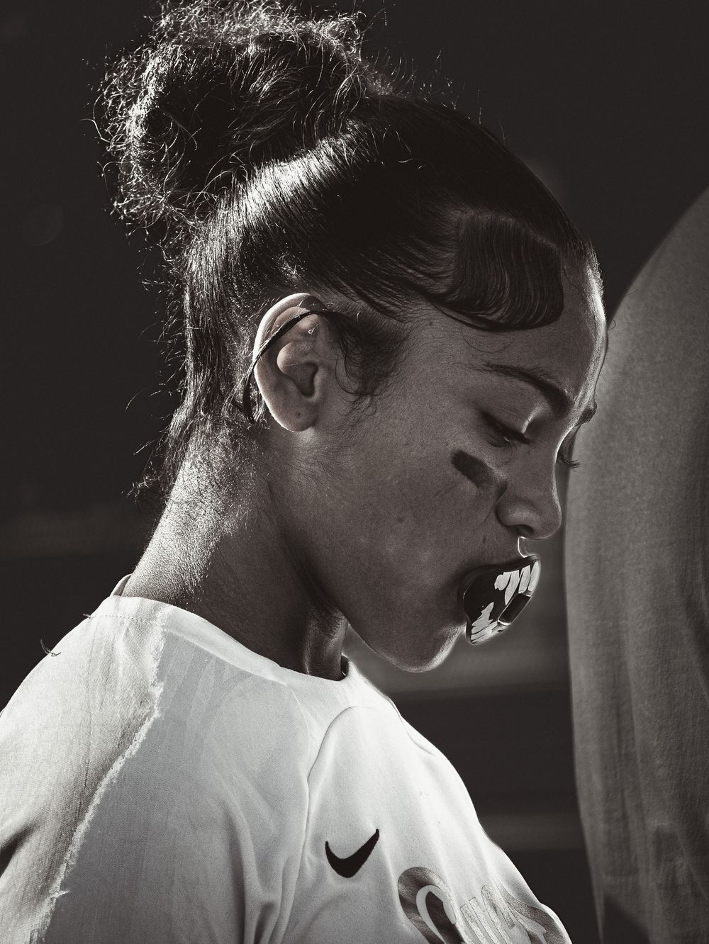 A black and white portrait shows a person in a Nike shirt with hair in a bun looking downward.