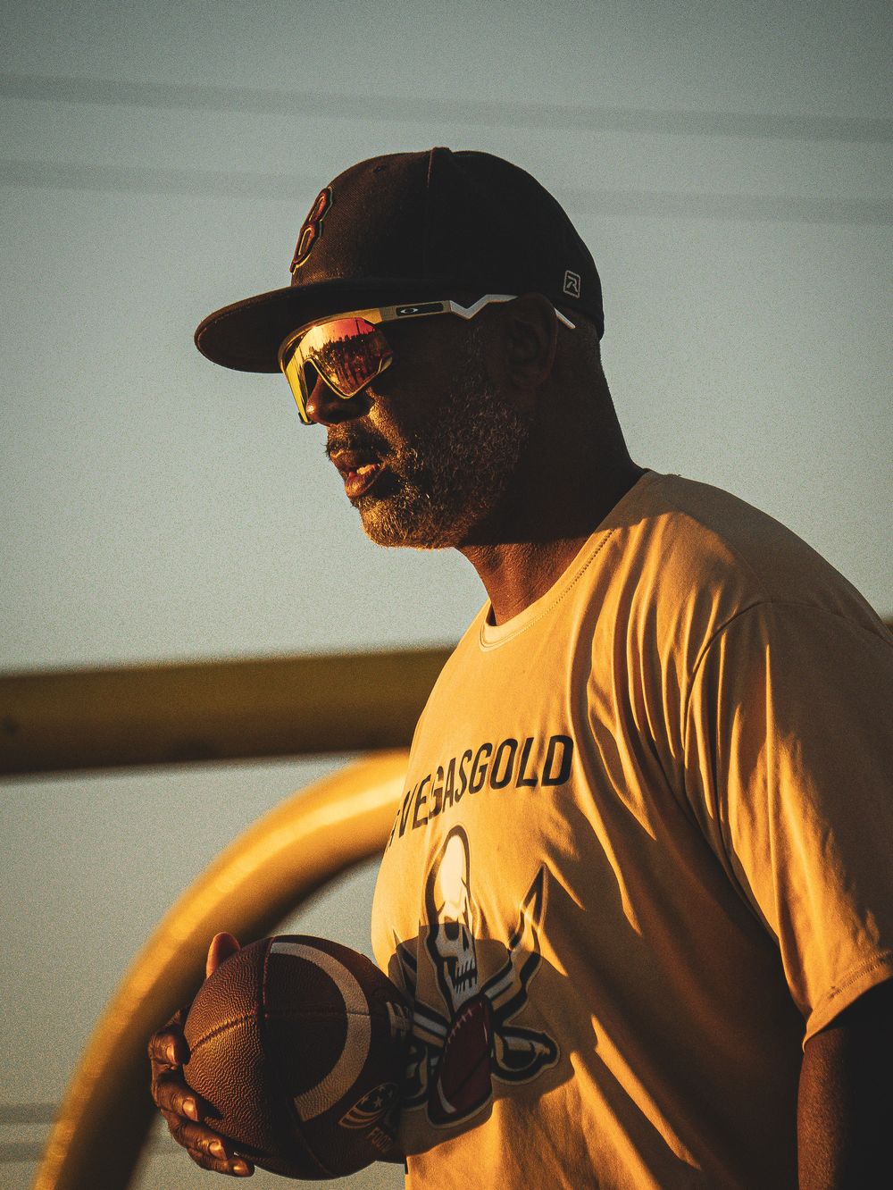 Coach in baseball cap holds football during golden hour lighting.