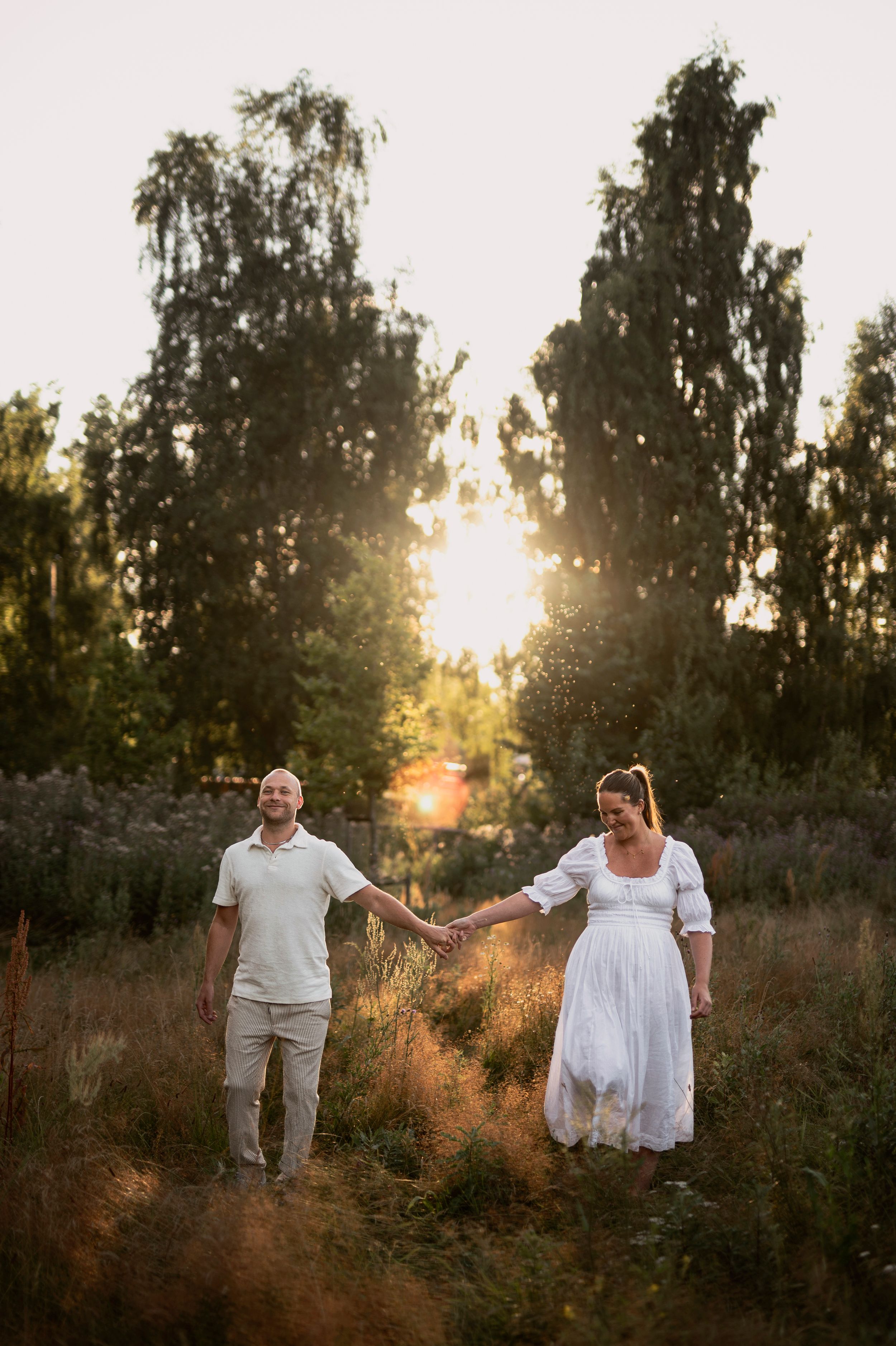 Par håller varandra i handen på en äng i golden hour, med björkar som silhuetter mot solnedgången.