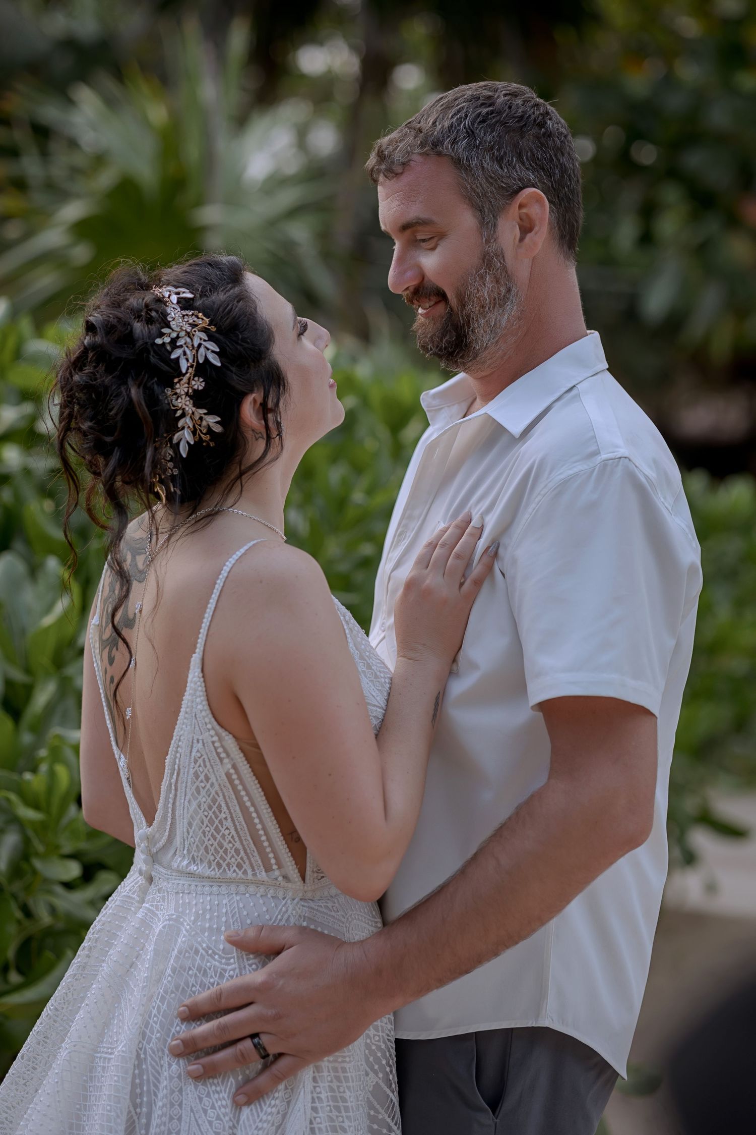 A romantic wedding couple embraces outdoors with lush greenery in the background while sharing an intimate moment.