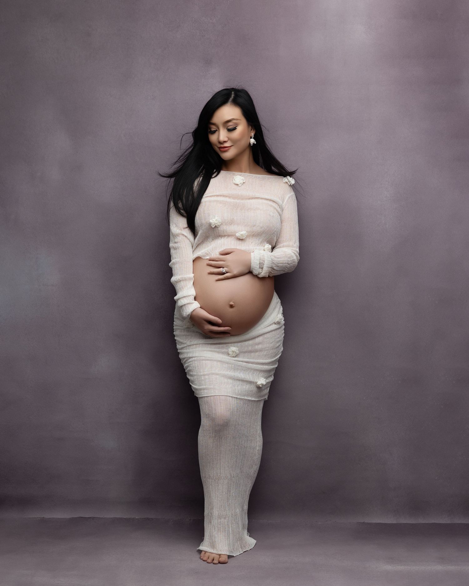 A maternity photo showing an expectant mother in a cream-colored dress standing against a gray backdrop.