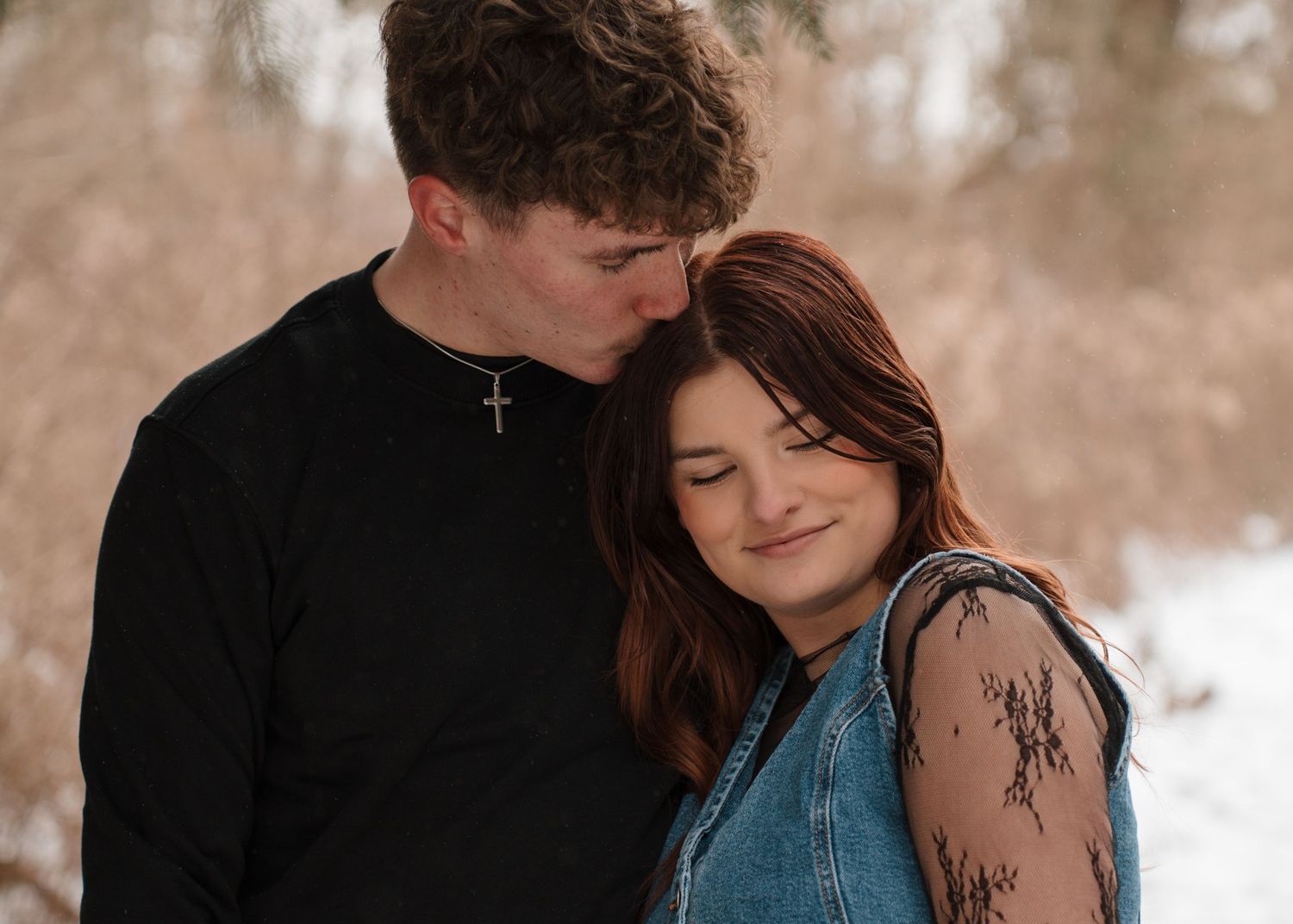 A couple shares a playful piggyback ride moment along a snowy winter path surrounded by bare trees.