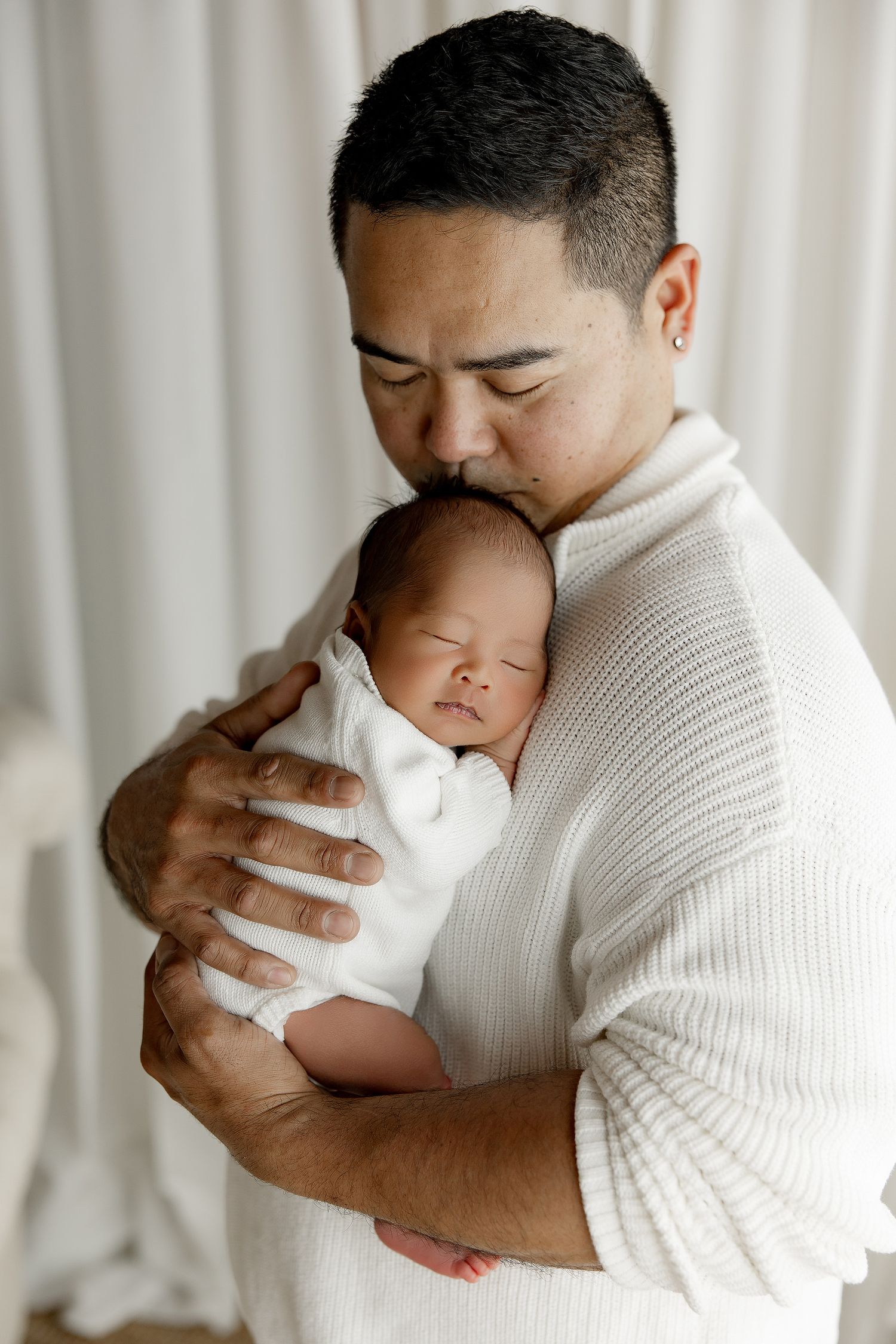A tender moment captured between parent and sleeping newborn baby in white against light curtained background.