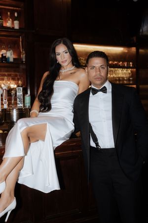 Elegant couple poses at a dimly lit bar, with her in a white dress and him in a black tuxedo with bow tie.