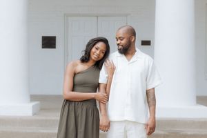A happy couple stands together outdoors, wearing casual summer outfits in olive green and white against a white building.