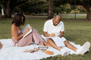 A group studies and takes notes while sitting together on a white blanket in a grassy park area.