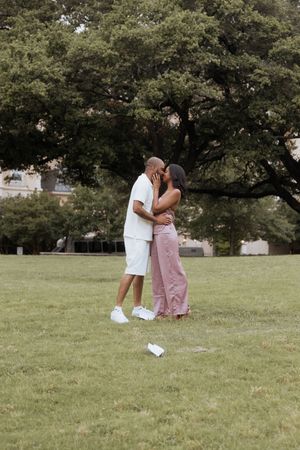 A couple shares a tender moment on a grassy lawn surrounded by lush green trees on a sunny day.