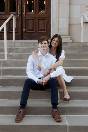 A couple sits together on concrete steps wearing coordinating white shirts in a casual engagement photo series.