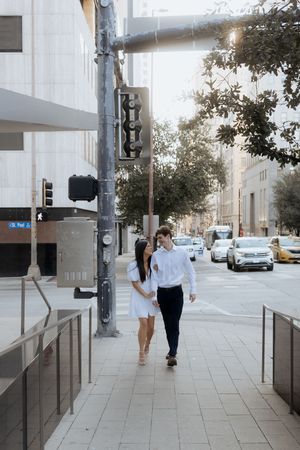 A couple walks down a modern city sidewalk on a sunny afternoon with parked cars and buildings visible in the background.