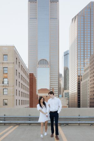 Two people pose for photos in front of modern glass skyscrapers and buildings in an urban setting.